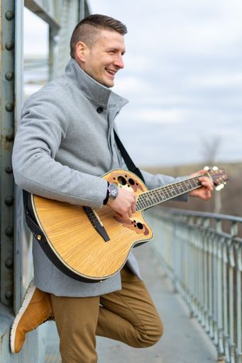 A vertical shot of a Caucasian white man with a gray coat playing the guitar on the bridge
