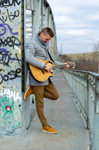 A vertical shot of a Caucasian white man with a gray coat playing the guitar on the bridge