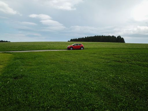 A red car driving through the green fields against a cloudy sky