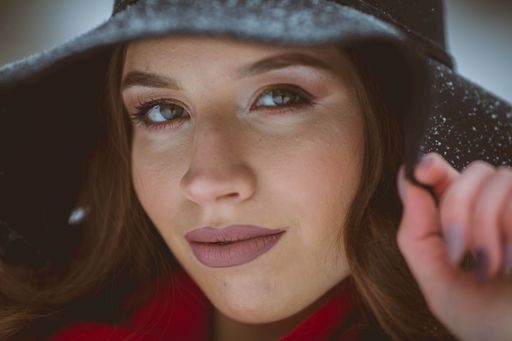 runette girl with perfect makeup holding her hat in a snowy forest while looking at the camera