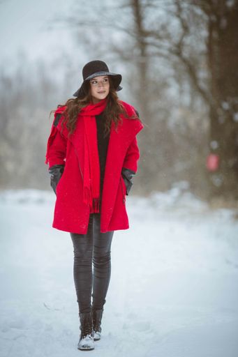 A full body shot of a young brunette girl wearing a stylish red coat and a hat walking in a snowy forest