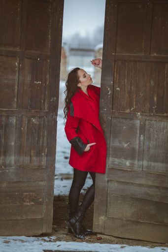 A full body shot of a young brunette girl wearing a stylish red coat standing between old wooden doors