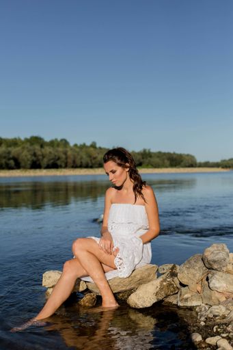 A beautiful white woman wearing a white dress and sitting down on the rock with her feet in the water