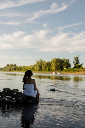 A beautiful white woman wearing a white dress and sitting down on the rock with her feet in the water