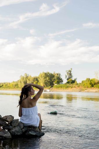 A beautiful white woman wearing a white dress and sitting down on the rock with her feet in the water
