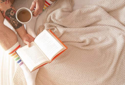 A top view of a tattooed man in rainbow socks drinking coffee and reading a book in bed in the morning