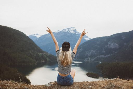 A behind shot of a young blond girl sitting on the cliff enjoying the view of the mountains and lake
