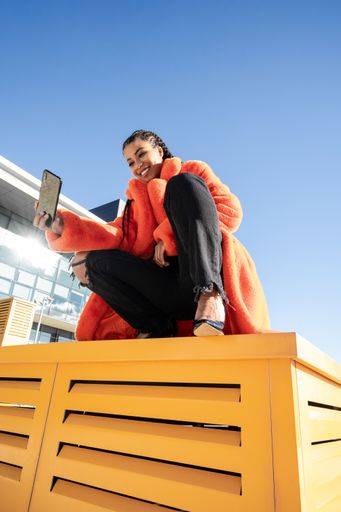 A vertical shot of a black female with a red coat sitting on a yellow surface and taking a selfie