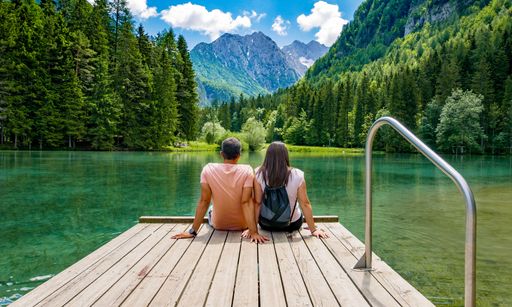 Rear view of young female sitting on wooden deck by beautiful lake in mountains. Green, spring, flower crown, outdoors. Zgornje Jezersko, Slovenia