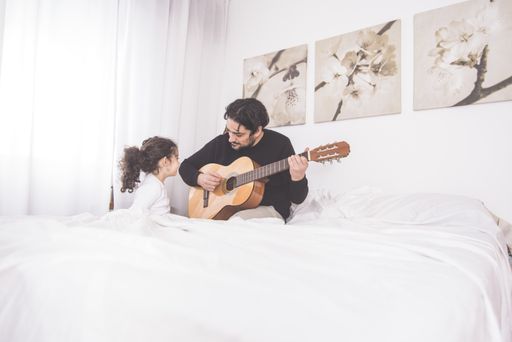 A father teaching his young daughter to play guitar sitting on a bed