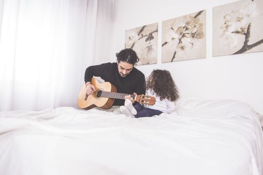 A father teaching his young daughter to play guitar sitting on a bed