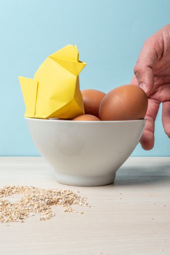 A vertical shot of a male hand and eggs with a yellow hen origami on blue background