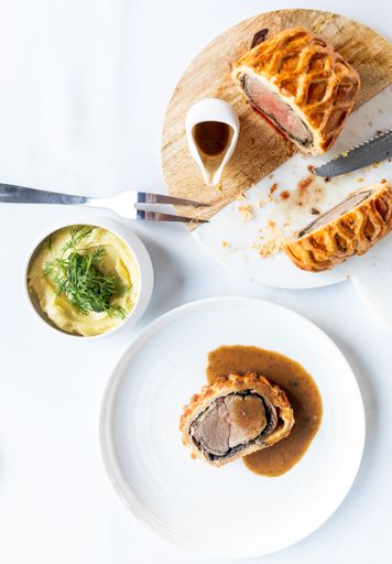 beef Wellington on a plate with mash potatoes and sauce, nicely plated on a white table cloth