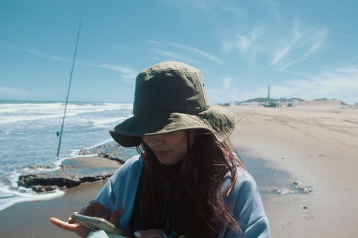 A young woman stands on a beach, smiling as she holds a phone in her right hand