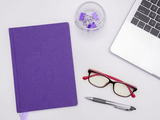 An organized workspace featuring a purple notebook, pen, glasses and a camputer