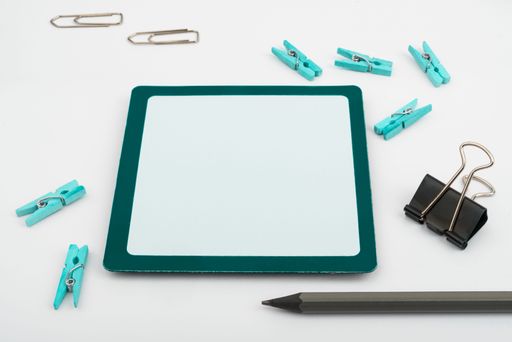 An assortment of multicolored paper clips scattered and a tablet with copy space in the middle atop a blank white sheet of paper