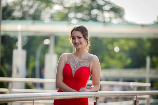A portrait of a Caucasian woman wearing a long red dress with jewellery and leaning on handrails in a park