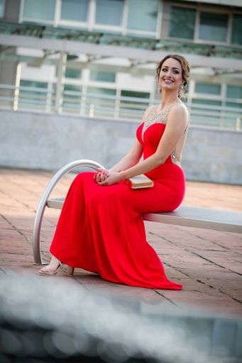 A shallow focus of a young Caucasian woman wearing a long red dress and sitting on a bench in a park