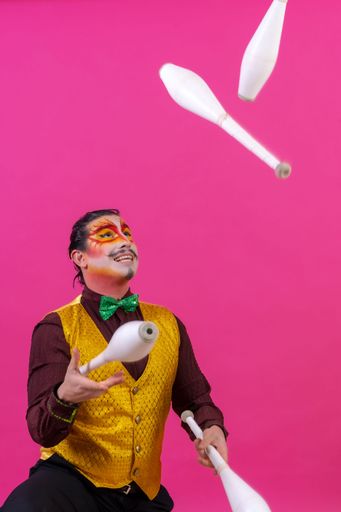 A cheerful circus performer skillfully juggling a number of clubs isolated on pink background
