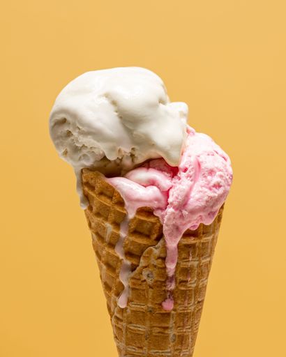 A melting ice cream in front of a yellow background. Vanilla and strawberry ice cream dripping down the cone.