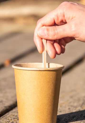 A male hand stirring a stick on a brown paper cup on a wooden surface