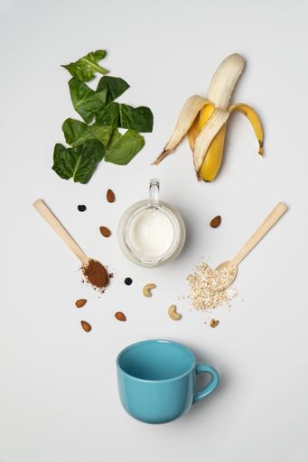 A top view of a white table with a mug, banana, greens, a pitcher with milk and other ingreidents