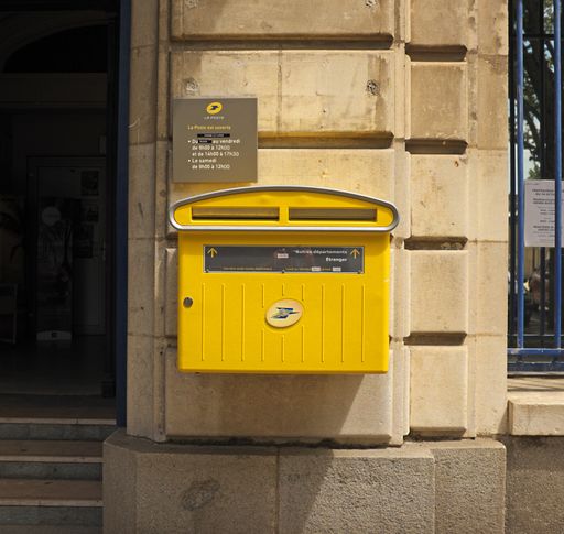 Yellow French mail box fixed to outside wall of a post office, next to entrance. Also visible is sign showing opening hours of post office.