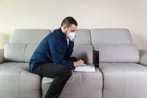 A male entrepreneur sitting on a couch wearing a protective mask working and talking on the phone