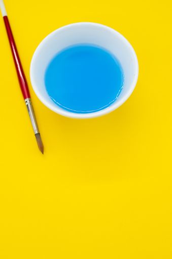 A vertical shot of a paint rinsing bowl with blue-stained water and a paintbrush on a yellow surface