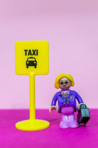 Playtive blond woman figurine with suitcase standing by a yellow board and waiting for a ordered taxi in a pink background.