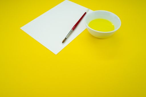 A top view of yellow liquid watercolor paint on a bowl with paper and paintbrush isolated on yellow background