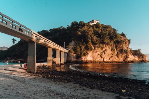 A beach surrounded by the sea and rocks covered in greenery with a bridge in Brazil