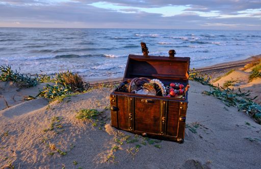 An open treasure chest filled with gold and jewelry on a beach at sunset