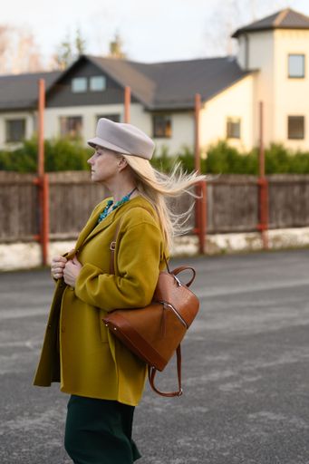 A young blonde woman wearing a mustard yellow jacket and a green skirt is accessorizing her look with a stylish hat