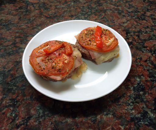 closeup shot of a homemade hamburger or ground meat burger with cheese, ham, tomato, red bell pepper and oregano in a plate on dark granite background