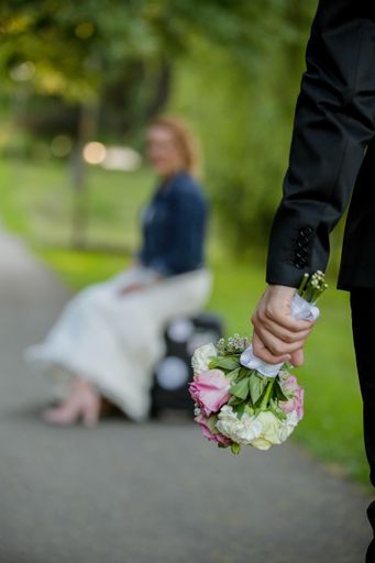 A vertical shot of the groom holding a flower bouquet with the bride sitting on a bench on the blurry background
