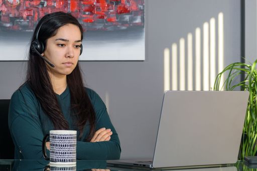 Annoyed South Asian woman having a business call in headphones at a desk, working on laptop in home office