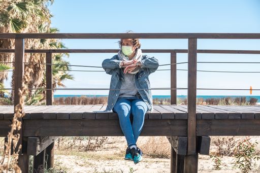 A closeup shot of a Spanish woman wearing a mask and sitting on a wooden bridge on a sunny day