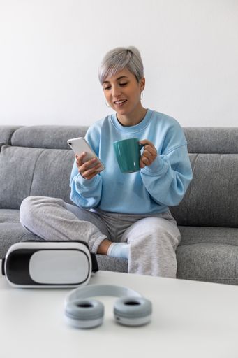 A young woman with short platinum blonde hair sitting on a sofa and drinking coffee while using her phone