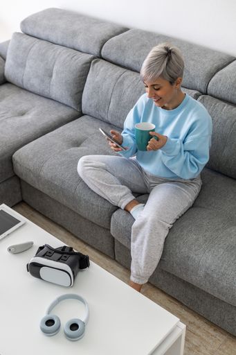 A young woman with short platinum blonde hair sitting on a sofa and drinking coffee while using her phone