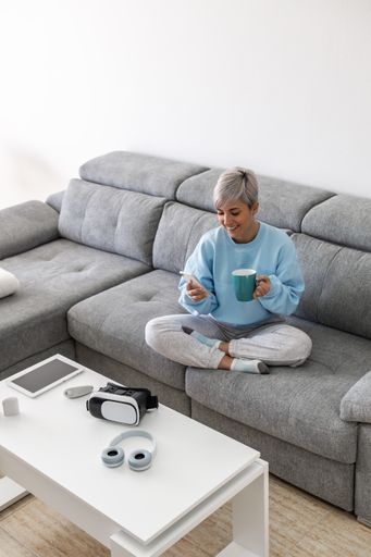 A young woman with short blonde hair sitting on a sofa and drinking coffee while using her phone