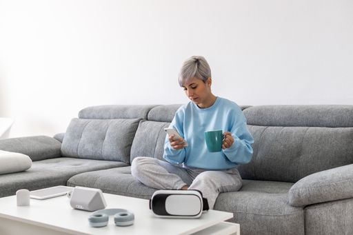 A young woman with short blonde hair sitting on a sofa and drinking coffee while using her phone