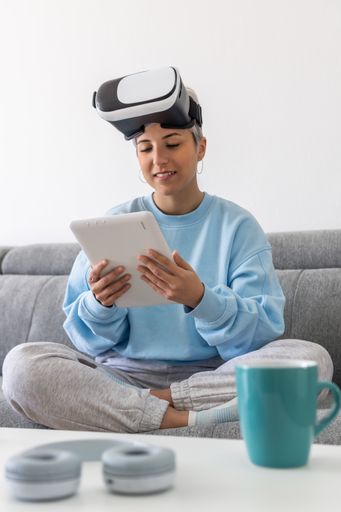 An adult woman using a VR headset and a tablet while sitting on a sofa