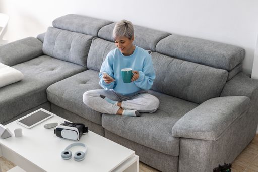 A young woman with short blonde hair sitting on a sofa and drinking coffee while using her phone