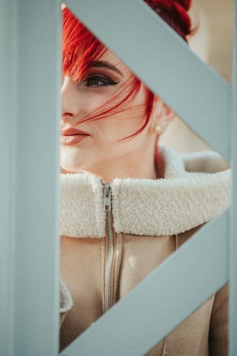 A portrait of a young red-haired girl with bangs behind a metal fence looking away on a windy winter day