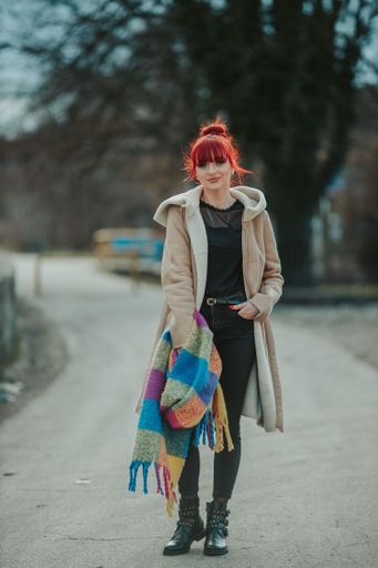 A full body shot of a young sexy red-haired woman with bangs strolling in the city holding a colorful scarf with a warm winter coat