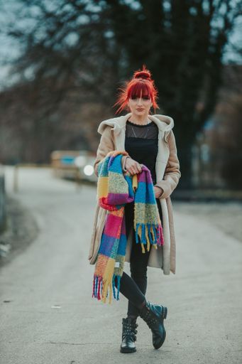 A full body shot of a young sexy red-haired woman with bangs strolling in the city holding a colorful scarf with a warm winter coat