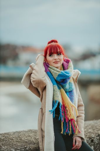 A portrait of a young sexy red-haired woman with bangs sitting on a stone wearing a colorful scarf with a warm winter coat
