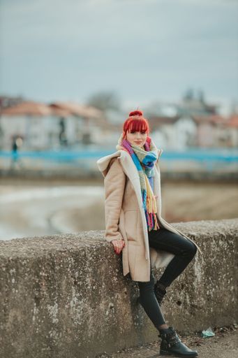A confident sexy red-haired woman with bangs sitting on a stone wearing a colorful scarf with a warm winter coat