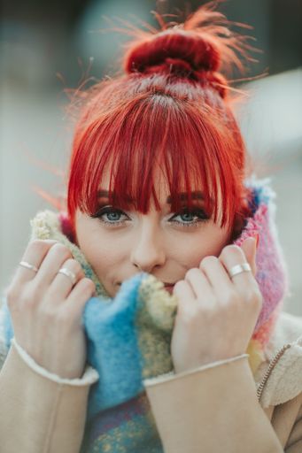 A portrait of a young red-haired girl with bangs holding her warm colorful scarf looking at the camera
